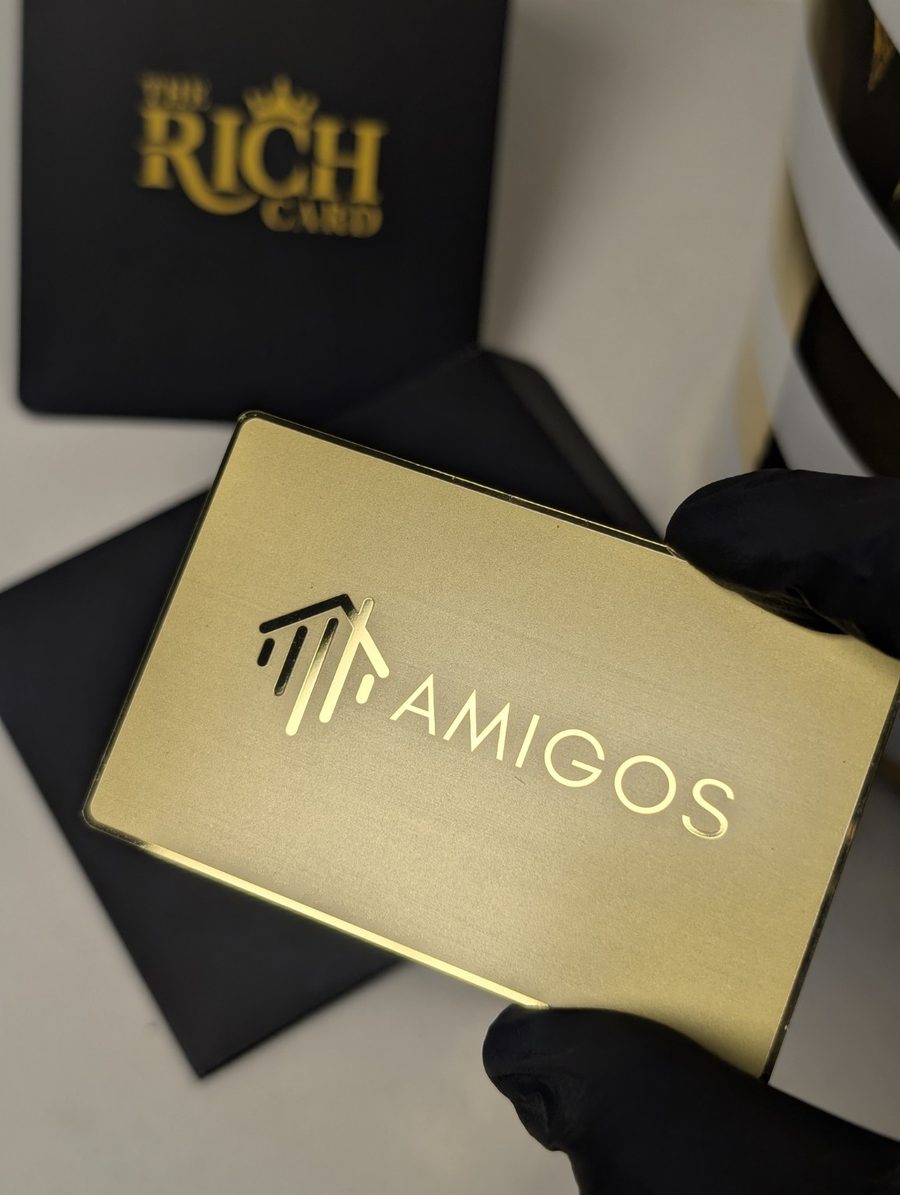 Gold-tone AMIGOS metal card with engraved logo, photographed with The Rich Card packaging